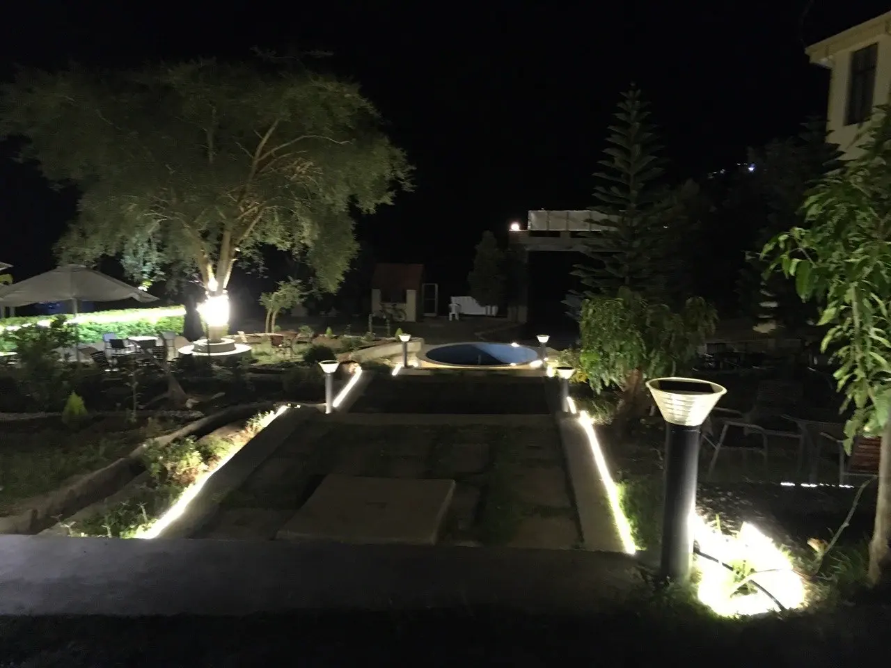 Garden path edged with lights at night, with trees and a low fountain beyond